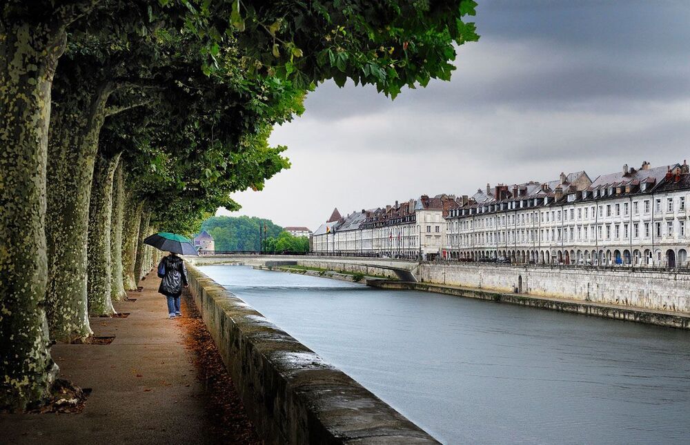A rainy day in the city of Besan&ccedil;on at the Quai Vauban. Besan&ccedil;on is a city in the east of France not far away from the Swiss border. The lady is walking past the Platane trees and at the other side of the Doubs river there are the old houses with small shops under the arcades.
