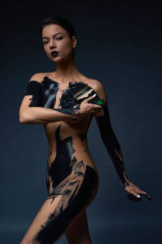 sensual slim young naked woman standing in dark studio and paint black color 