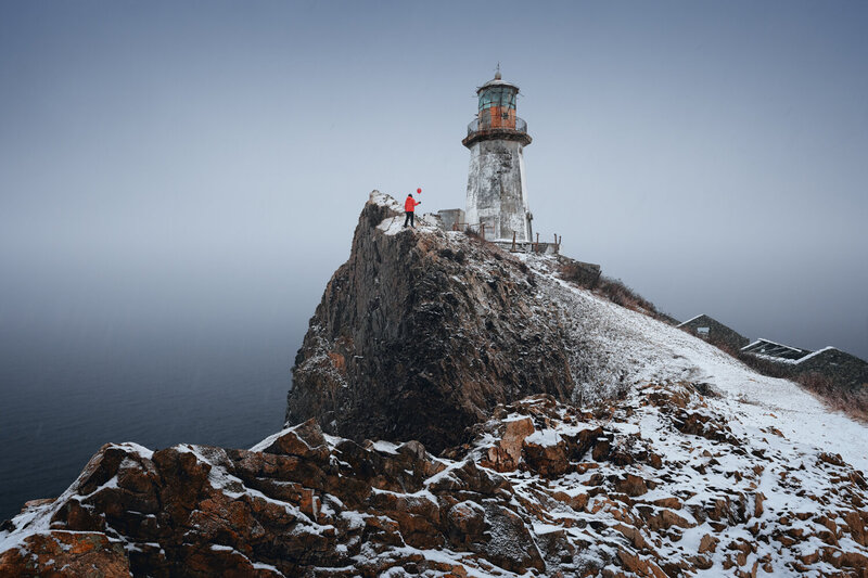 lighthouse, cliff, rocks, snow, winter, person, sea, isolated, nature, seascape, coastal, solitary figure, cold, atmospheric, moody, gray sky, rugged terrain, outdoor, пейзаж, маяк, зима, приморье, снег Один на вершине фото превью